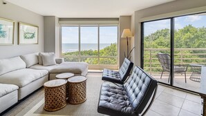 Un salon moderne comprenant un canapé, deux fauteuils, une table basse et un lampadaire. De grandes fenêtres offrent une vue sur l’océan et la végétation environnante.
