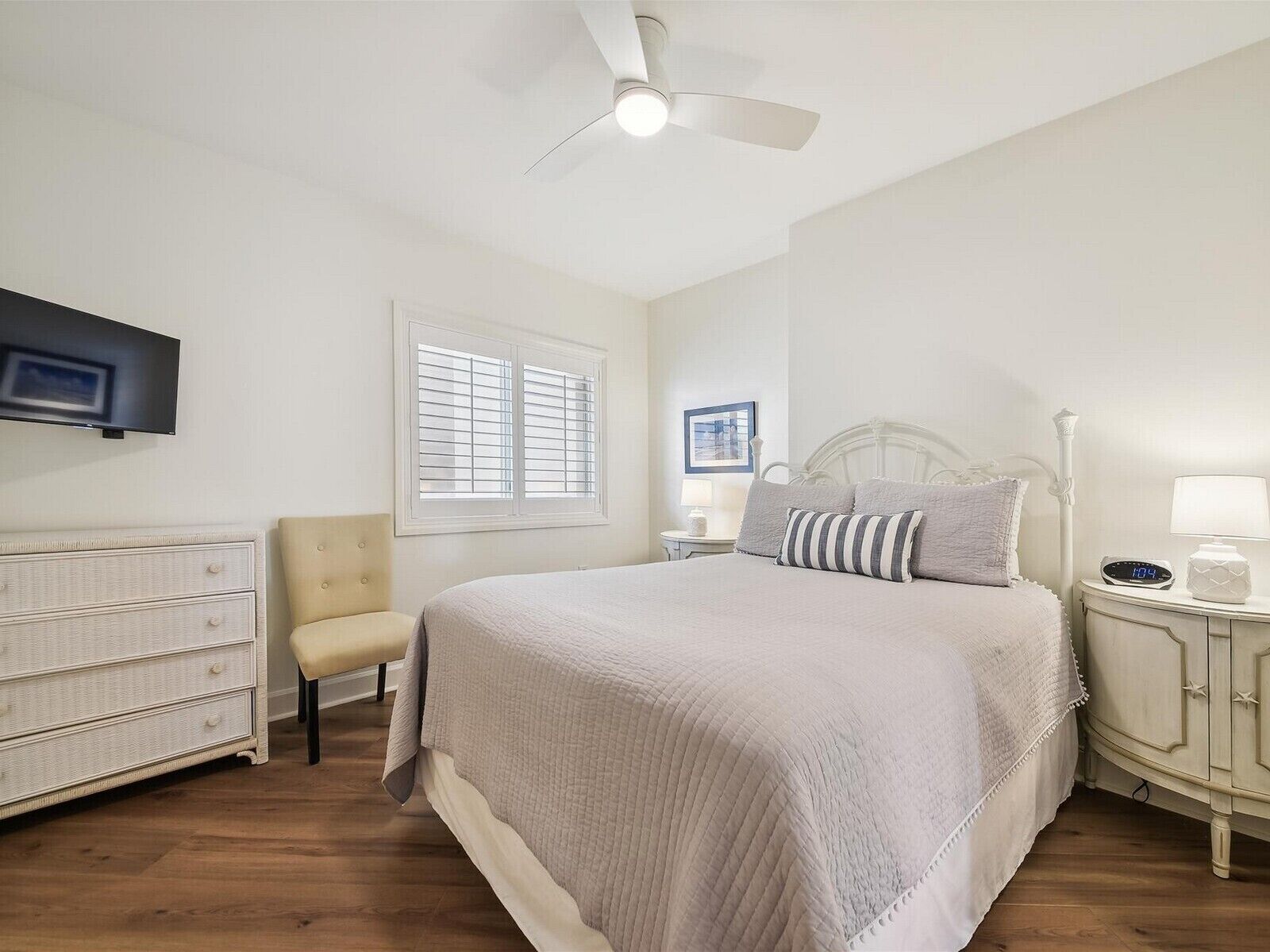 Un dormitorio con cama, una silla, cómoda, mesita de noche, lámpara, reloj, ventilador de techo y ventana con persianas.