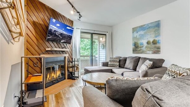 A modern living room with a fireplace, a large sofa, and a flat-screen TV mounted on a wooden wall. - Lovely 2 BDR Condo - Close to Skiing & Main St! (Breckenridge)