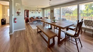 A spacious living room with a large wooden dining table, multiple chairs, and a sofa. There is a fireplace, a large window with blinds, and a ceiling fan. - Perch - LFP View Home: Cozy 4BR Home, Lake Washington View Deck, Backyard (Lake Forest Park)