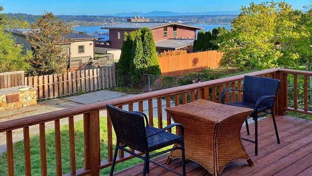 A wooden deck with two wicker chairs and a table, overlooking a body of water and distant buildings. - Perch - LFP View Home: Cozy 4BR Home, Lake Washington View Deck, Backyard (Lake Forest Park)