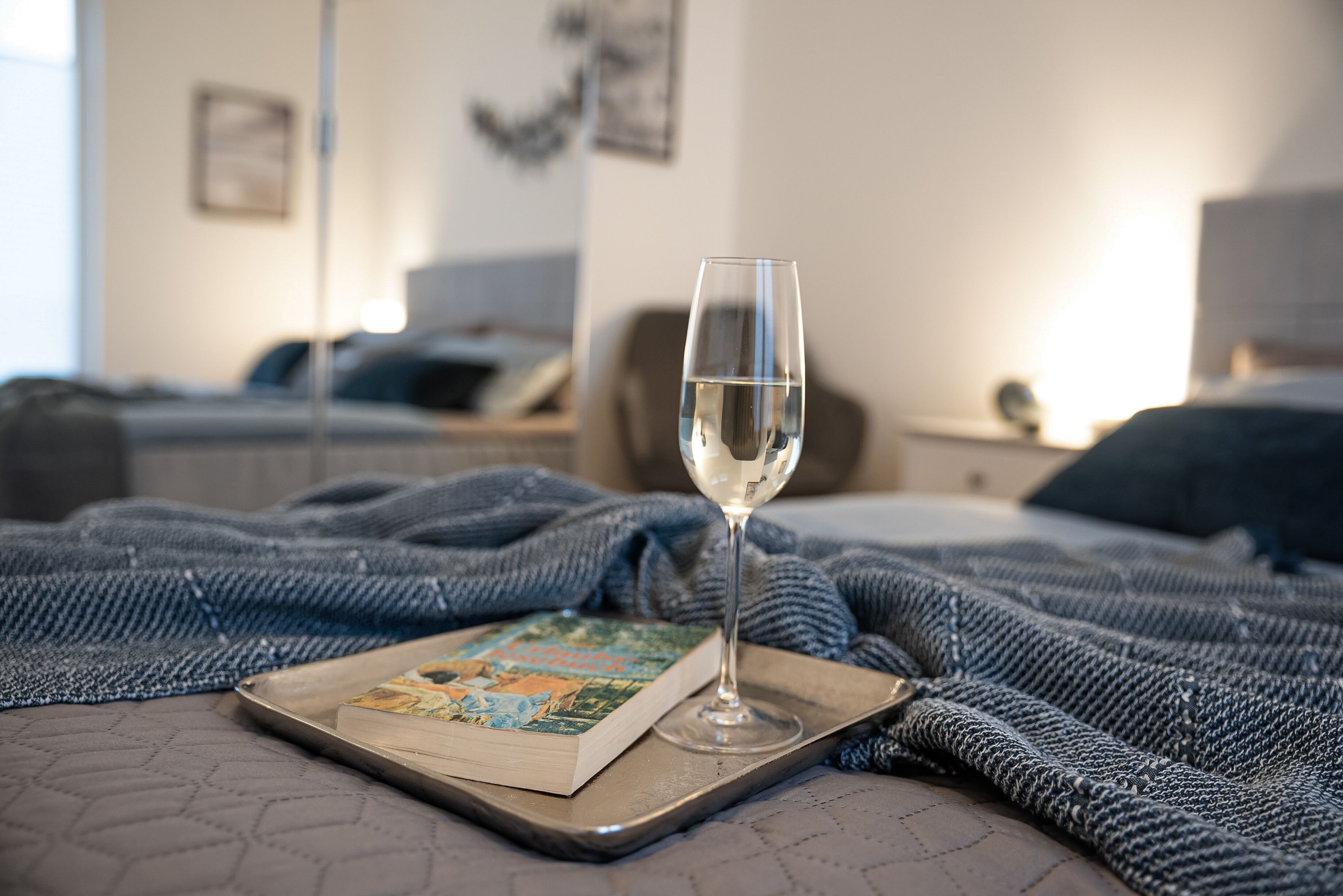 A glass of water on a tray with a book on a bed, surrounded by a blue knitted blanket.