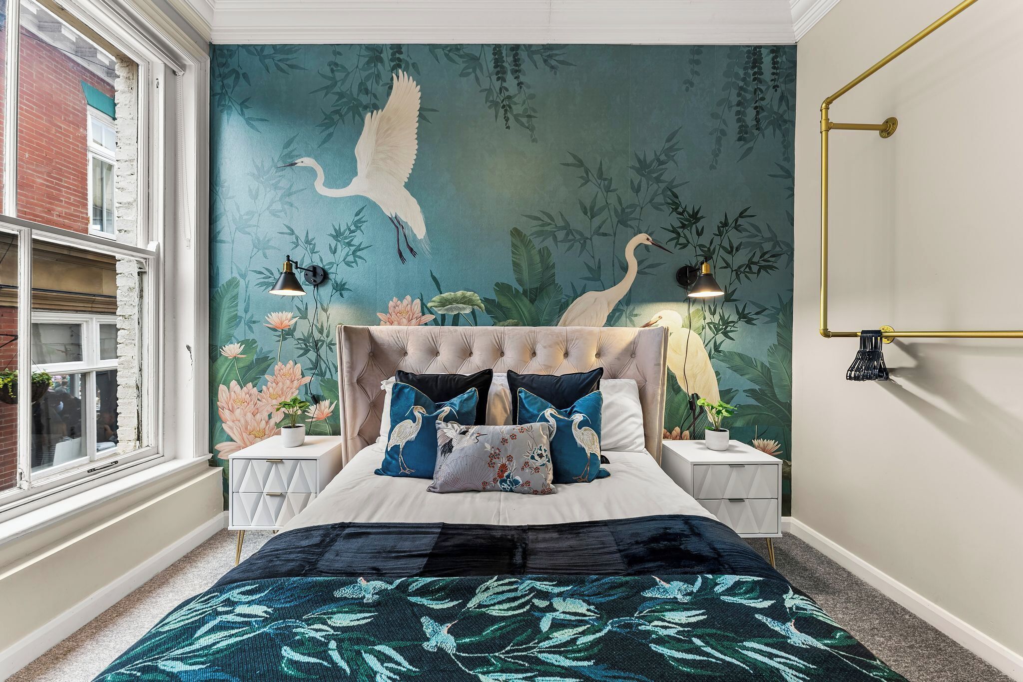 Quarto com cabeceira estofada, cama com almofadas de motivos florais e de pássaros, uma janela grande e um mural de bambu e aves na parede.