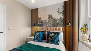 Ein Schlafzimmer mit einer gepolsterten Kopfstütze, einem Bett mit türkis-goldfarbenen Kissen, einer grünen Decke, einer Wandpaneele aus Holz und einem Fenster mit einer Pflanze.