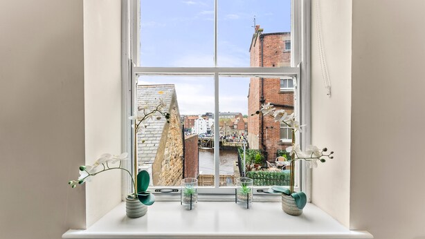 Ein Fenster mit vier Blumentöpfen auf einer weißen Ablage, mit Blick auf eine Stadtlandschaft mit Ziegelgebäuden und blauem Himmel.