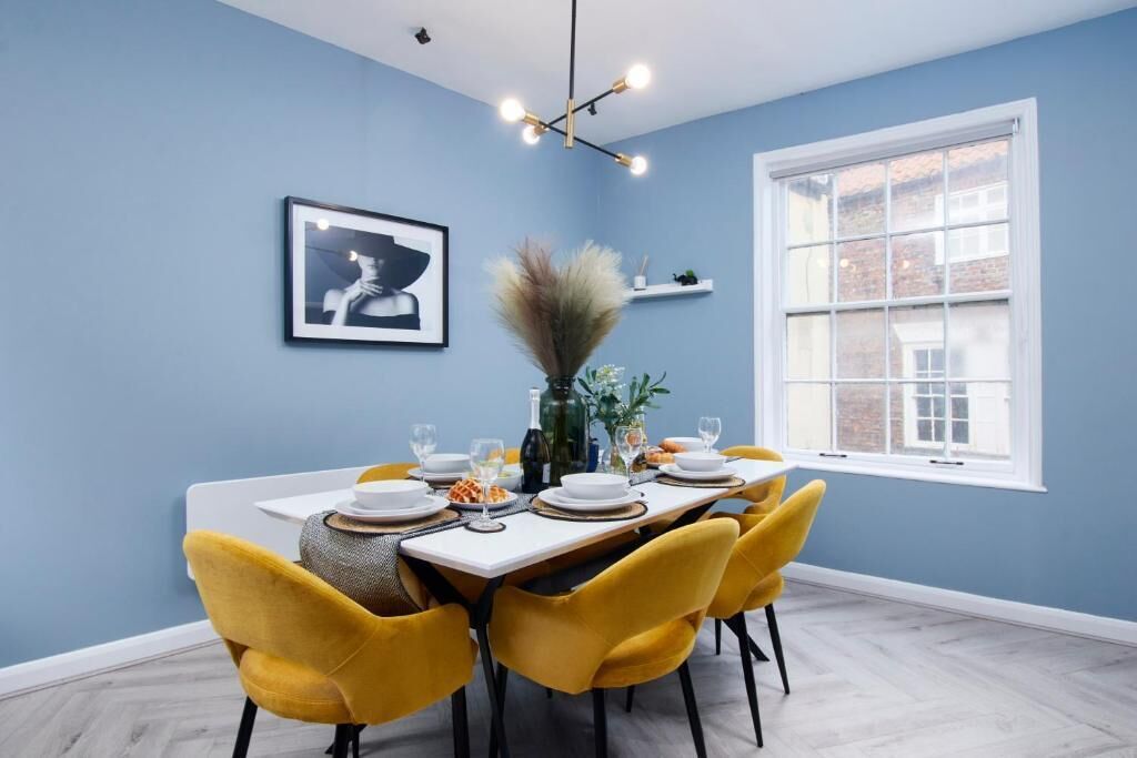 Een eetgedeelte met een tafel voor vier personen, gele stoelen, een kroonluchter, een lijst met een afbeelding aan de muur en een raam met uitzicht op gebouwen.