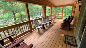 A covered porch with wooden benches and chairs, large windows, and a view of a forested area.