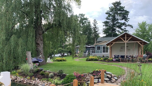 A wooden deck over a pond with a rope barrier, surrounded by lush greenery and a house with a porch.