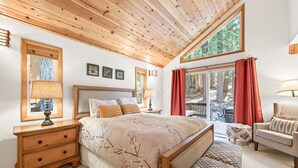 A bedroom with a wooden ceiling, a large bed, a nightstand with a lamp, a chair, and a view of trees through a window. - Bear's Lair by Tahoe Getaways - Luxury Cabin w Hot Tub & HOA Access (Truckee)