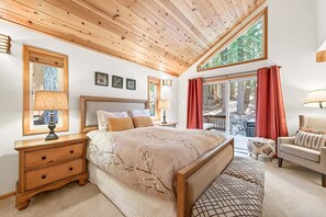 A bedroom with a wooden ceiling, a large bed, a nightstand with a lamp, a chair, and a view of trees through a window.