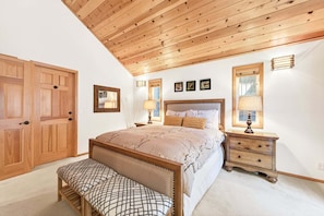 A bedroom with a wooden ceiling, a bed with a headboard, bedside tables, a bench, and a door.