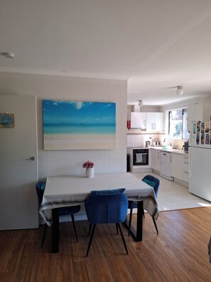 Una cuina moderna amb armaris blancs, una zona de dinar amb taula i cadires, i un quadre gran amb tema de platja a la paret.