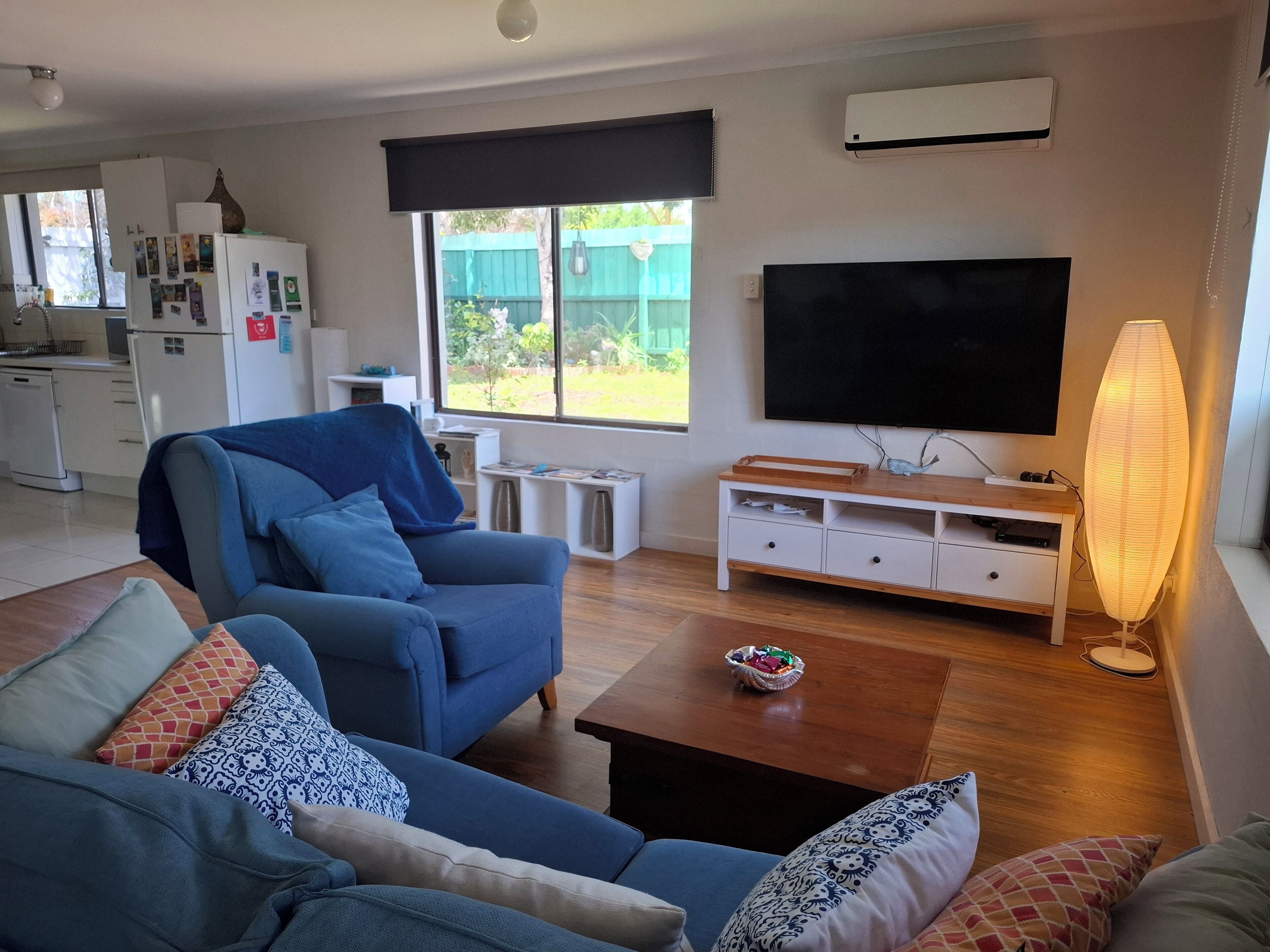 Un salón con un sofá azul, una mesa de centro, un televisor y una lámpara. Se ve una cocina al fondo con un refrigerador y una ventana.