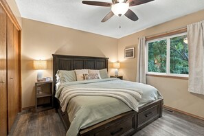 A bedroom with a bed, nightstands, a ceiling fan, a window with curtains, and a view of trees. - Family Retreat | Hot Tub, Fire Pit & Game Room (Sioux Falls)
