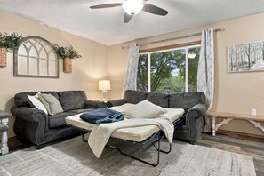 A living room with a sofa, a coffee table, a lamp, and a window with curtains. - Family Retreat | Hot Tub, Fire Pit & Game Room (Sioux Falls)
