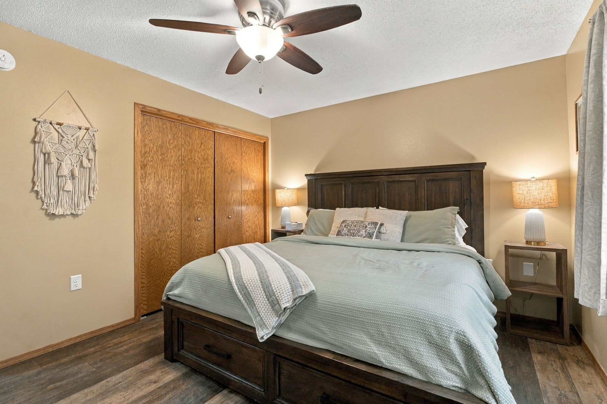 A bedroom with a bed, a ceiling fan, a wardrobe, and a nightstand with a lamp.