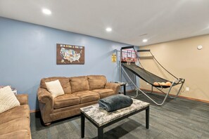A living room with a brown sofa, a small table, a hammock, and a wall-mounted basketball hoop. - Family Retreat | Hot Tub, Fire Pit & Game Room (Sioux Falls)