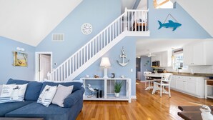 Un vaste espace de vie comprenant un canapé bleu, un plancher en bois, un escalier blanc et une cuisine avec des meubles blancs.