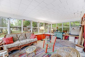 A sunroom with a sofa, chairs, a dining table, and a kitchen area with a refrigerator and cabinets.
