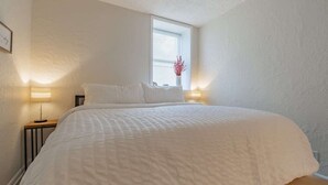 Quarto com cama grande, duas luminárias de cabeceira, janela com persianas e um vaso com flores.