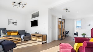 En moderne stue med en grå sofa, en rød lænestol og et træbord. Der er et fjernsyn monteret på væggen og en bogreol i baggrunden.