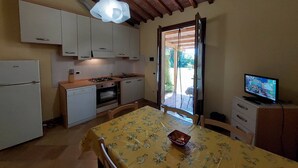A compact kitchen with white cabinets, a gas stove, a sink, and a dining table with a floral tablecloth. - Lovely flat with private covered terrace, pool, large shared garden. (Toscana)