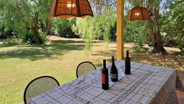 An outdoor dining area with a table set for two, surrounded by chairs, under a pergola with hanging lamps. - Lovely flat with private covered terrace, pool, large shared garden. (Toscana)