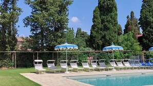 A poolside area with lounge chairs and umbrellas, surrounded by greenery and trees. - Lovely flat with private covered terrace, pool, large shared garden. (Toscana)