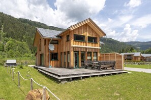 Eine Holzhütte mit einer Terrasse, umgeben von einer Wiese und Bergen im Hintergrund.