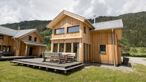 Uma cabana de madeira com varanda e área de estar externa, rodeada por uma encosta arborizada.