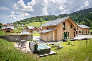 Un chalet en bois doté d’un bain à remous et d’un espace extérieur aménagé, entouré d’un paysage verdoyant luxuriant et bordé, au loin, par une colline boisée.