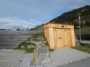 A wooden structure with a slanted roof, situated on a paved area with a stone and grass border, adjacent to a road and a mountain backdrop.