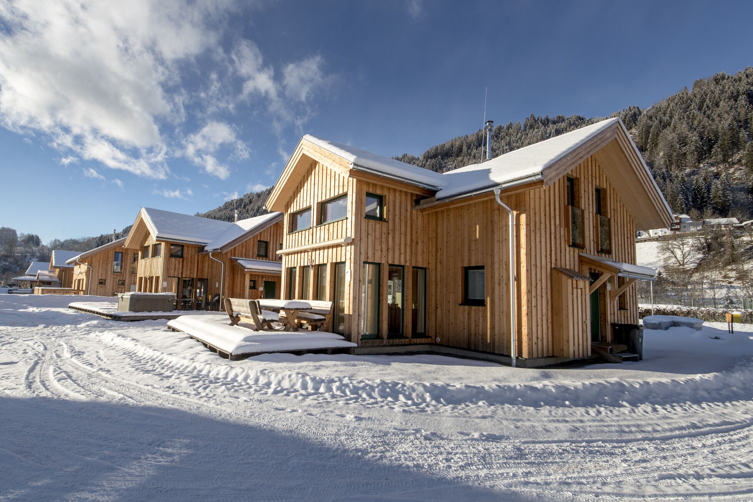 Un conjunto de cabañas de madera con techo nevado, un área de estar al aire libre techada y vista a una montaña boscosa en el fondo.
