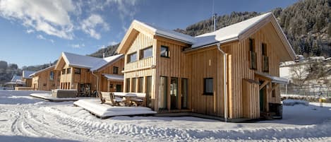 Un conjunto de cabañas de madera con techo nevado, un área de estar al aire libre techada y vista a una montaña boscosa en el fondo.