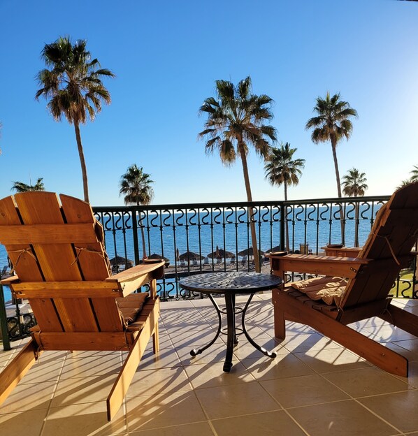 A terrace with wooden furniture, a round table, and a view of the sea with palm trees.