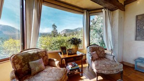 A room with two upholstered chairs, a wooden table, and a view of a mountainous landscape through large windows.