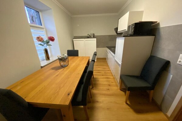 Een compacte keuken met een houten eettafel, zwarte stoelen en een klein raam met bloemen.