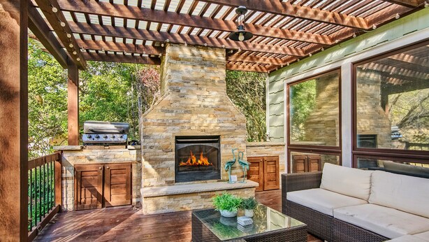 Een terras buiten met een stenen open haard, een barbecue, rieten meubels en een pergola.