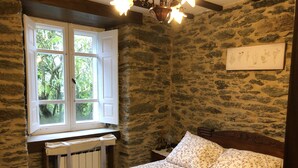 Ein Schlafzimmer mit Steinwand, einem Fenster mit Blick ins Grüne, einem Bett mit Blumenbettwäsche und einer Wandleuchte.