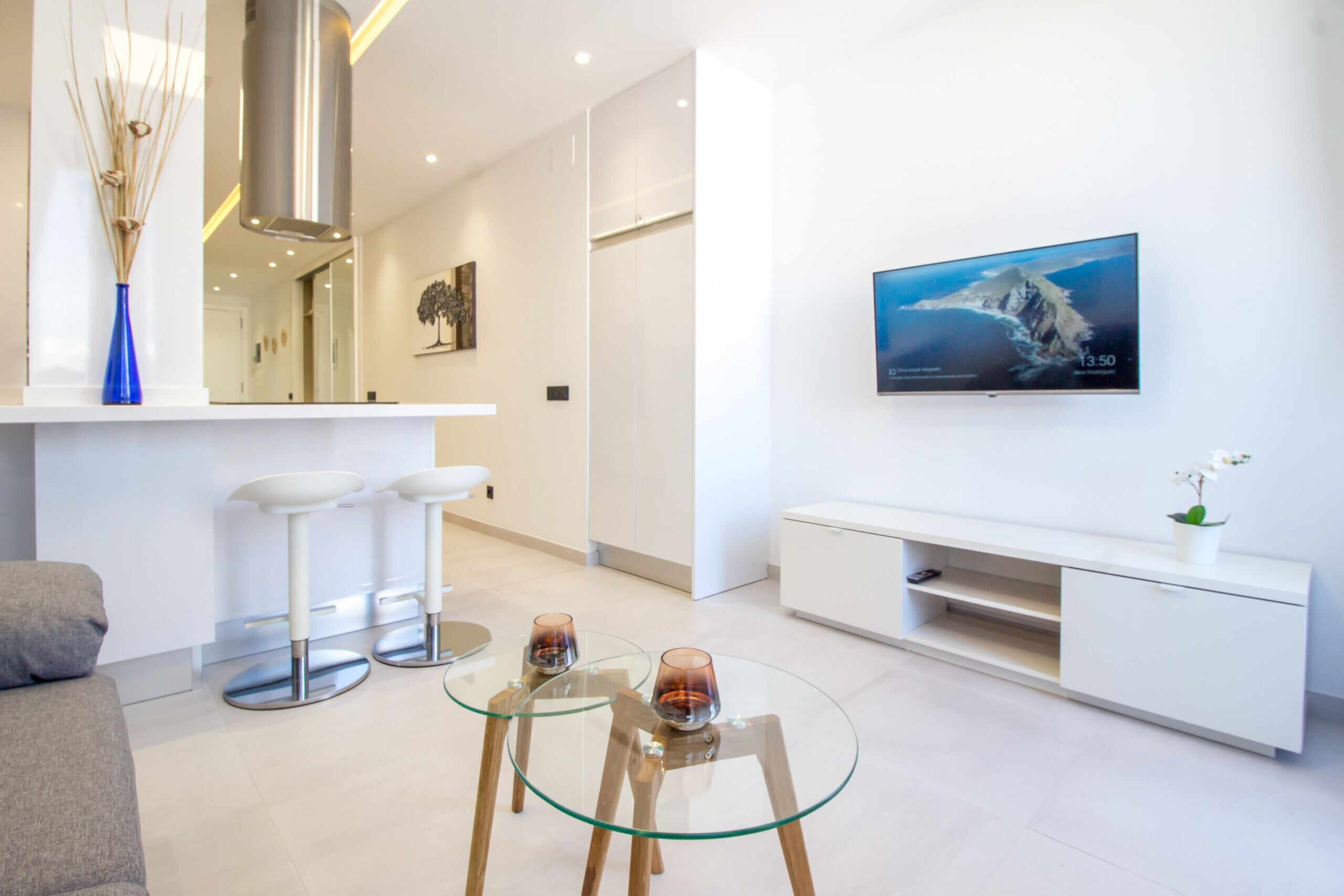 A modern living room with a glass coffee table, white sofas, a TV mounted on the wall, and a minimalist design.