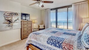 A bedroom with a large bed, a ceiling fan, a TV on a wooden dresser, a painting of a boat, and a view of the beach through a large window. - Oceanviews-MultiBalconies-Steps 2 Beach-Pool/HotTub-Gated Community-Kingston (Myrtle Beach)