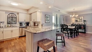 A modern kitchen with white cabinets, a marble island, and a dining area with dark wooden chairs and a chandelier. - Oceanviews-MultiBalconies-Steps 2 Beach-Pool/HotTub-Gated Community-Kingston (Myrtle Beach)