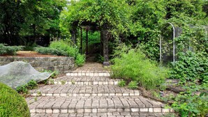 A stone pathway leading to a wooden gazebo surrounded by lush greenery and a metal fence.
