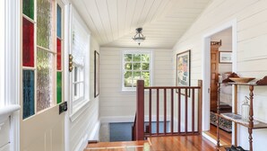 A hallway with wooden floors, white walls, and a wooden railing. There is a window with a view of greenery outside.