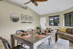A dining area with a long table, chairs, and a ceiling fan. There are framed pictures on the wall and a window with a view.