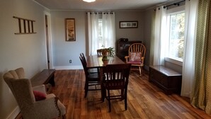 A dining area with a wooden table and chairs, a bookshelf, and a window with curtains.