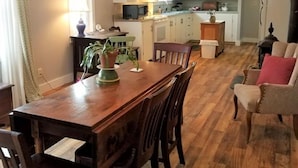 A dining area with a wooden table and chairs, a kitchen in the background, and a living room with a sofa and armchair.