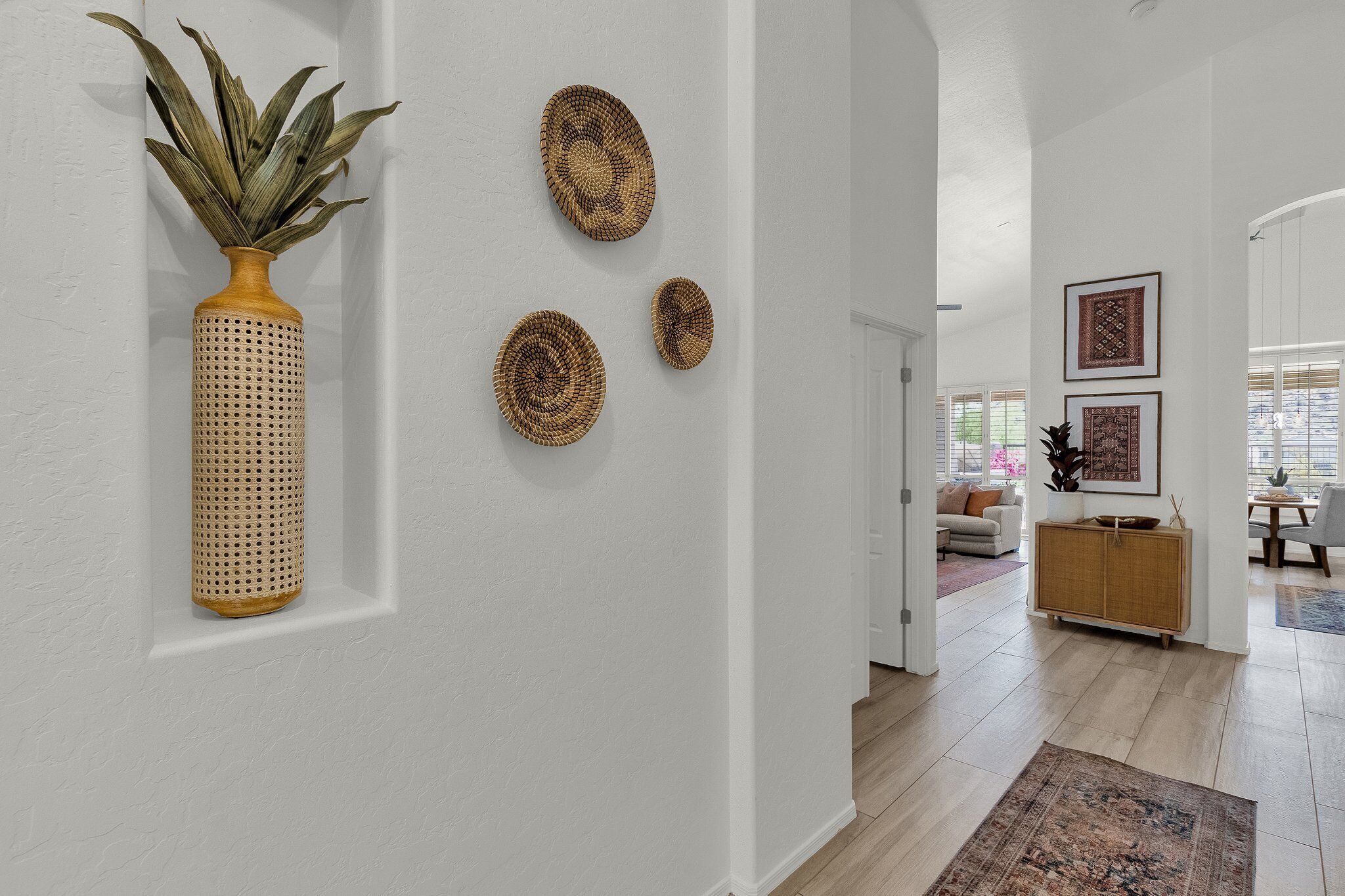 Un pasillo con un jarrón tejido y tres tapices, que conduce a un espacio de sala con un sofá y un mueble de madera.