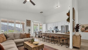 A spacious living room with a large sectional sofa, a wooden coffee table, and a kitchen area with white cabinets and stainless steel appliances. - Incredible golf course views with modern, southwest inspired decor (Mesa)
