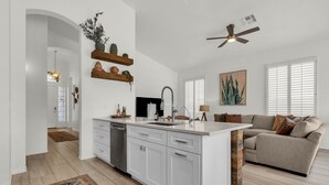 A modern kitchen with white cabinetry, a built-in dishwasher, and a central island. There is a ceiling fan, a window with blinds, and a living area with a sofa and a painting. - Incredible golf course views with modern, southwest inspired decor (Mesa)